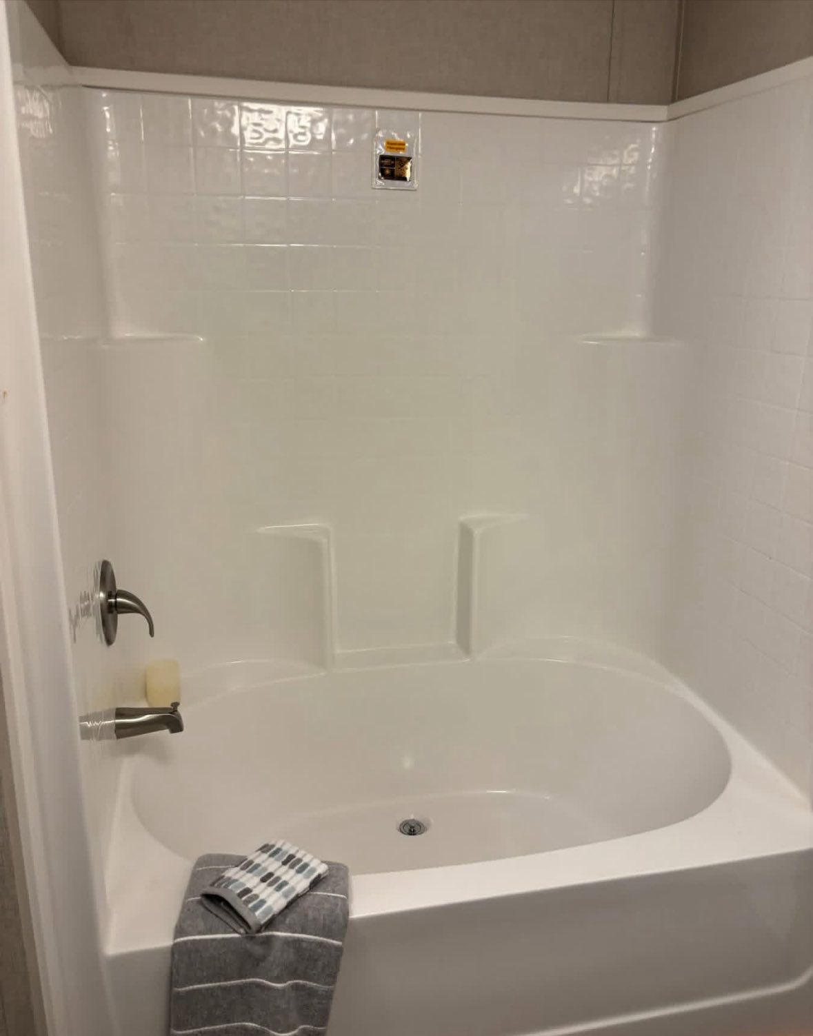 White bathtub with built-in shelf, shiny tile surround, and silver faucet. A small soap bar and folded gray-striped towel add a fresh touch.