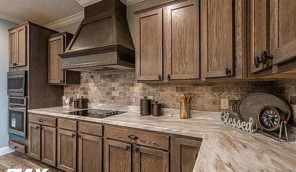 Warm, rustic kitchen with brown wooden cabinets, marble countertops, and stone backsplash. Items include jars, utensils, a clock, and a "Blessed" sign.