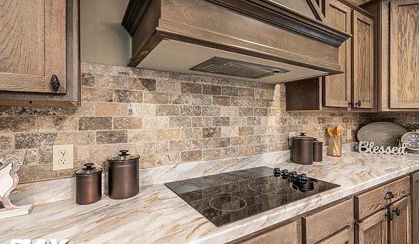 Warm kitchen with brown cabinetry, marble countertop, and brick backsplash. Includes a stovetop and "Blessed" decor, creating a cozy, rustic feel.