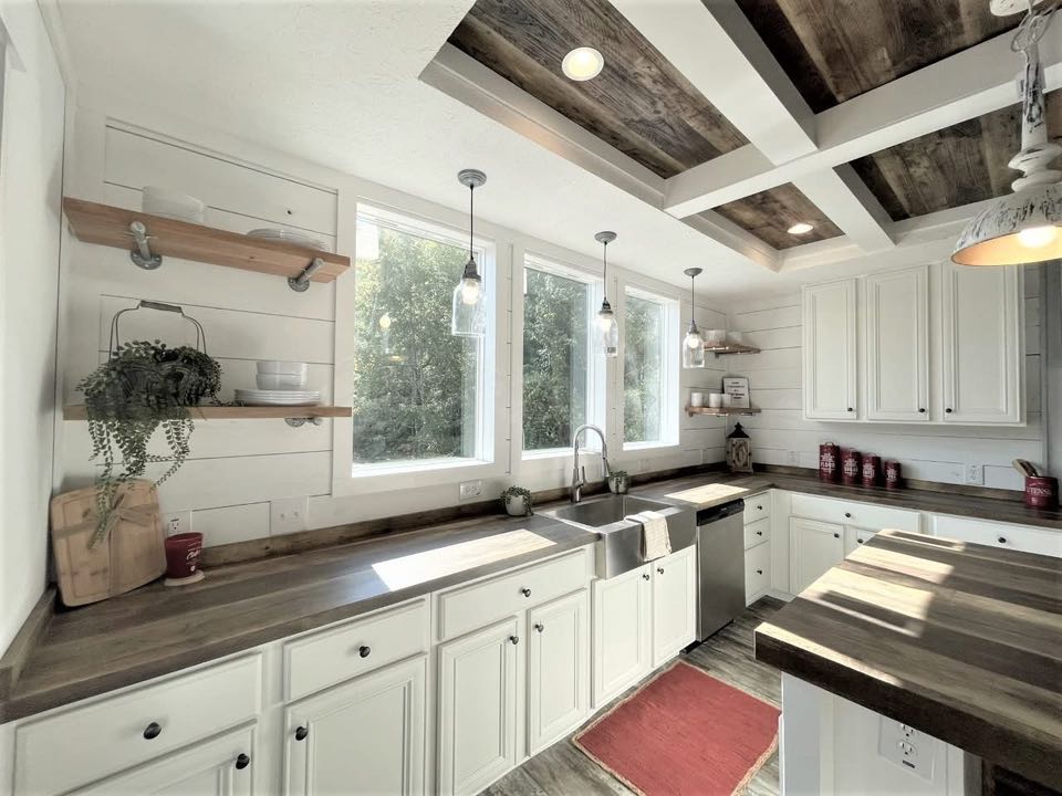 Bright farmhouse kitchen with white cabinets, rustic wood countertops, and a large apron sink. Natural light streams through three large windows.