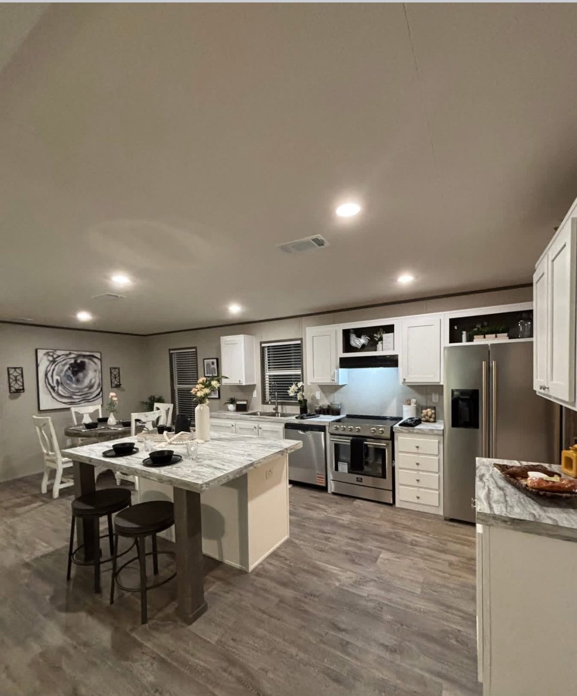 Modern kitchen with white cabinets, gray marble countertops, and stainless steel appliances. A breakfast bar with stools and a table are visible. Warm lighting adds a cozy feel.