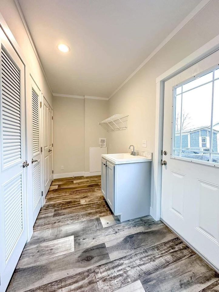 Bright laundry room with wood-style flooring and white walls. Features a utility sink, overhead drying rack, and storage closets. Glass door lets in natural light.