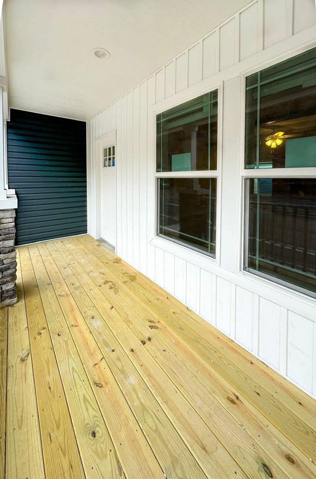 Wooden porch with light yellow planks, white walls, and large windows on the right. A small door and dark blue siding are at the far end.
