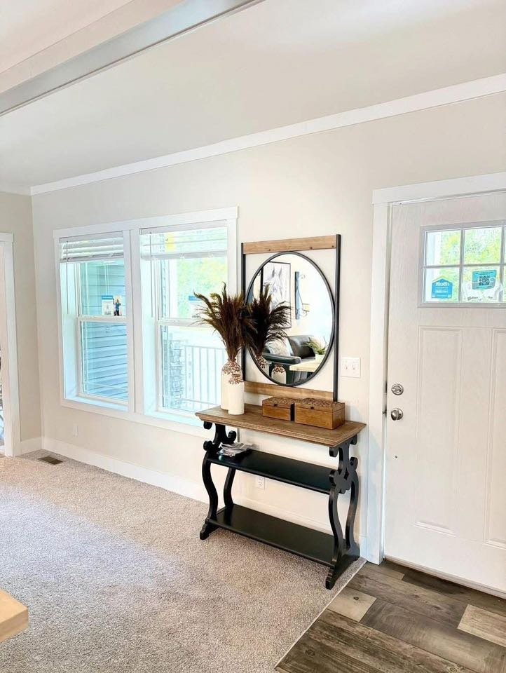 Bright entryway featuring a round mirror above a rustic wooden table with decorative vases and plants, near windows and a white door. Cozy and welcoming.