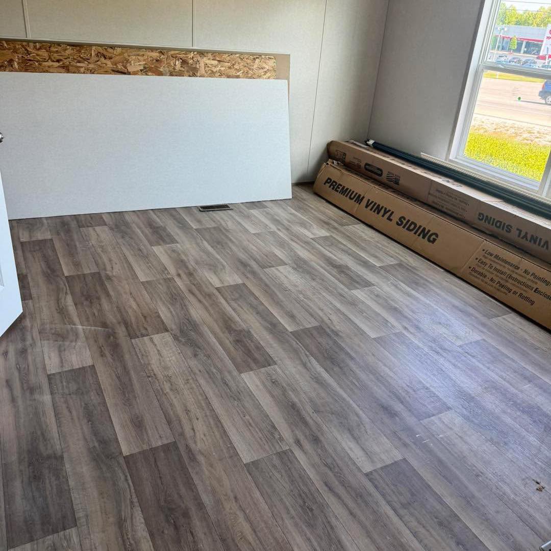 Room with light wood plank flooring, a white panel leaning against the wall, and boxes labeled "Premium Vinyl Siding" near a large window. Bright ambiance.