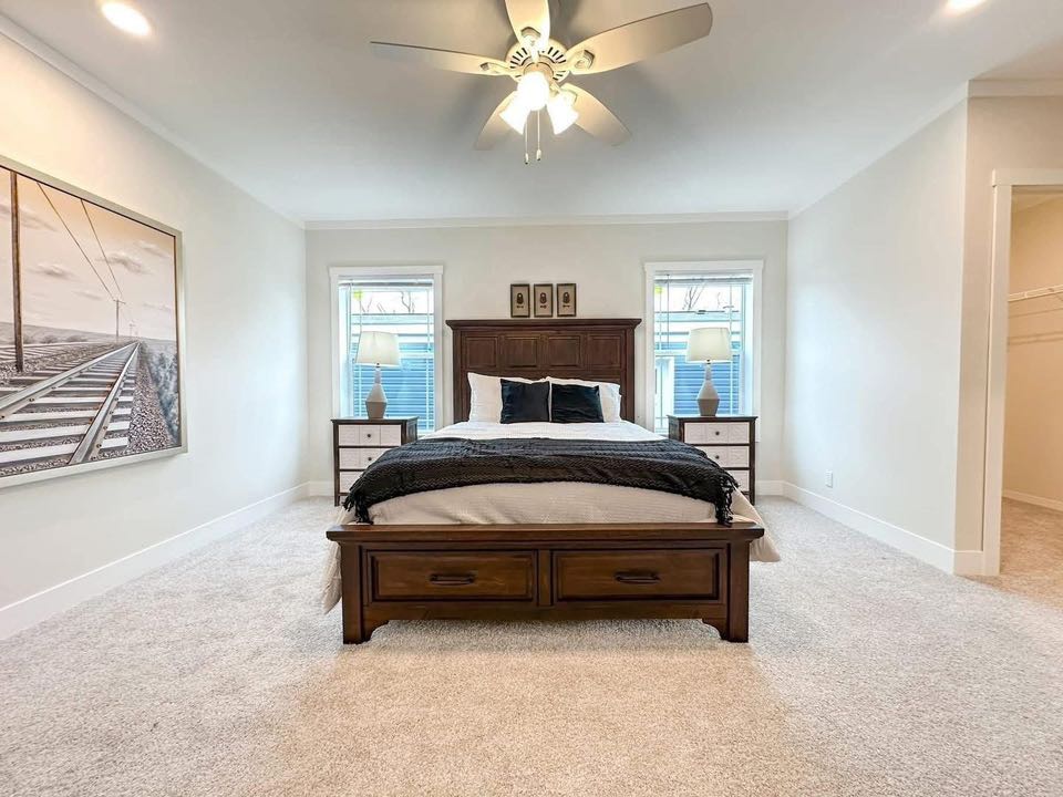A spacious bedroom with a wooden bed, white bedding, and dark pillows. Two windows with lamps flank the bed. A calm, minimalist tone is set with soft lighting and a ceiling fan. A large artwork of railroad tracks adorns the wall.