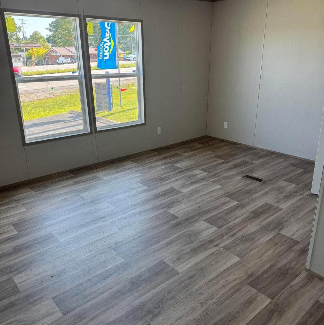 An empty room with light wood-patterned vinyl flooring and gray walls. Two large windows show a street view with green grass and a blue flag outside.