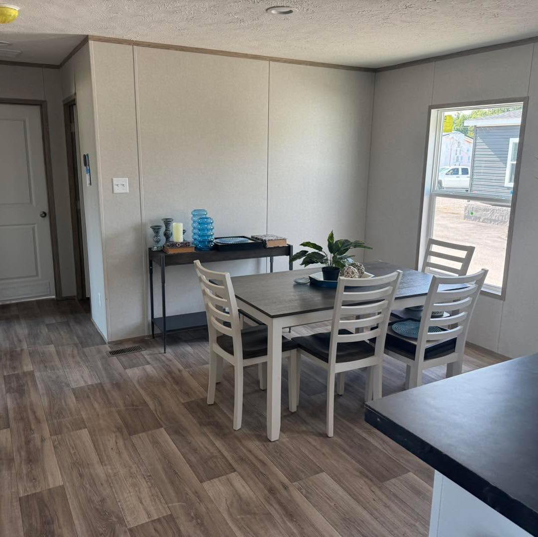 Minimalist dining area with wooden flooring features a table set for four with potted plant centerpiece. A console table with decor is against a plain wall. Natural light streams through a window, creating a peaceful ambiance.