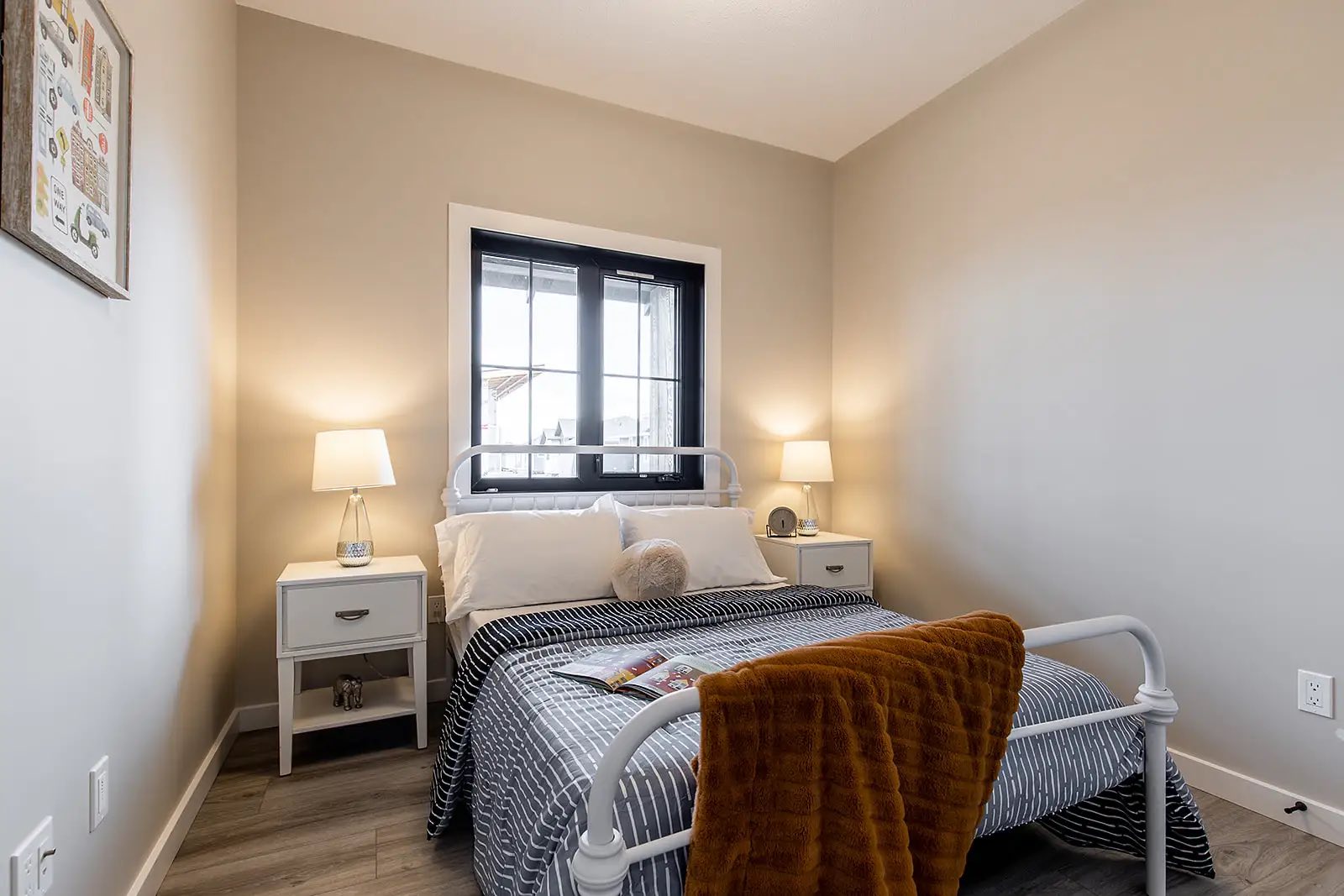 A cozy bedroom with a neatly made bed, featuring striped bedding and an orange throw. Soft lighting from two lamps on white side tables creates a warm and inviting atmosphere.