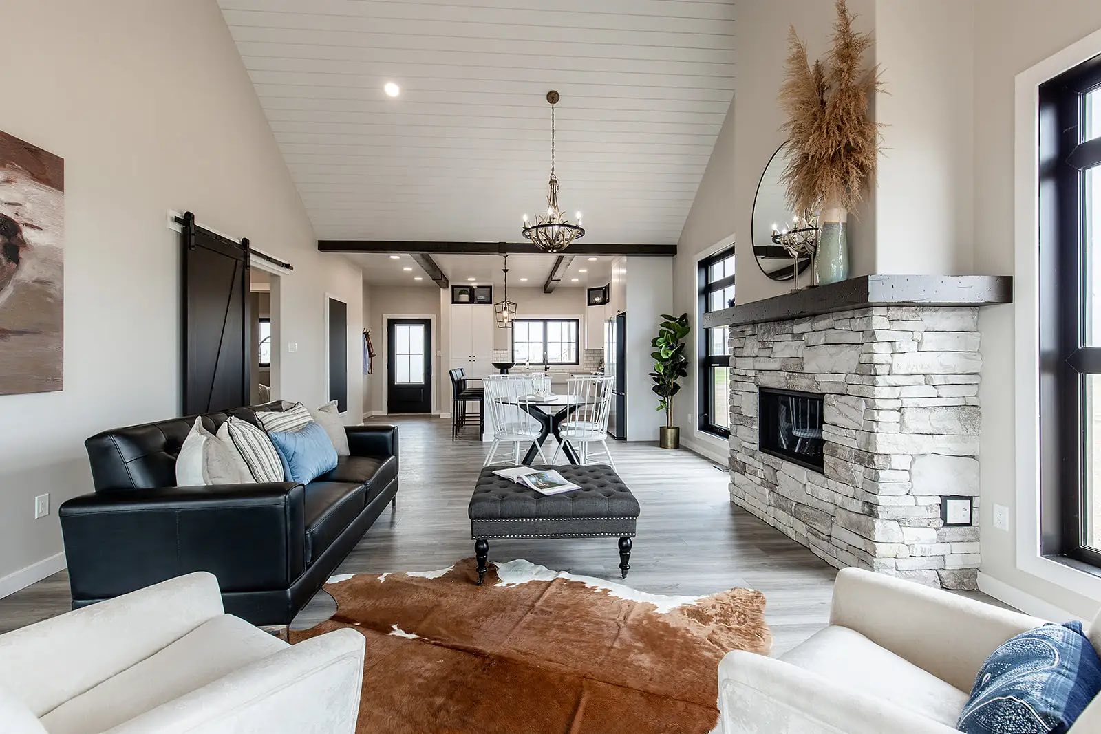 Spacious modern living room with vaulted ceiling, stone fireplace, and elegant chandeliers. Features black sofa, white armchairs, and dining area. Cozy and inviting.