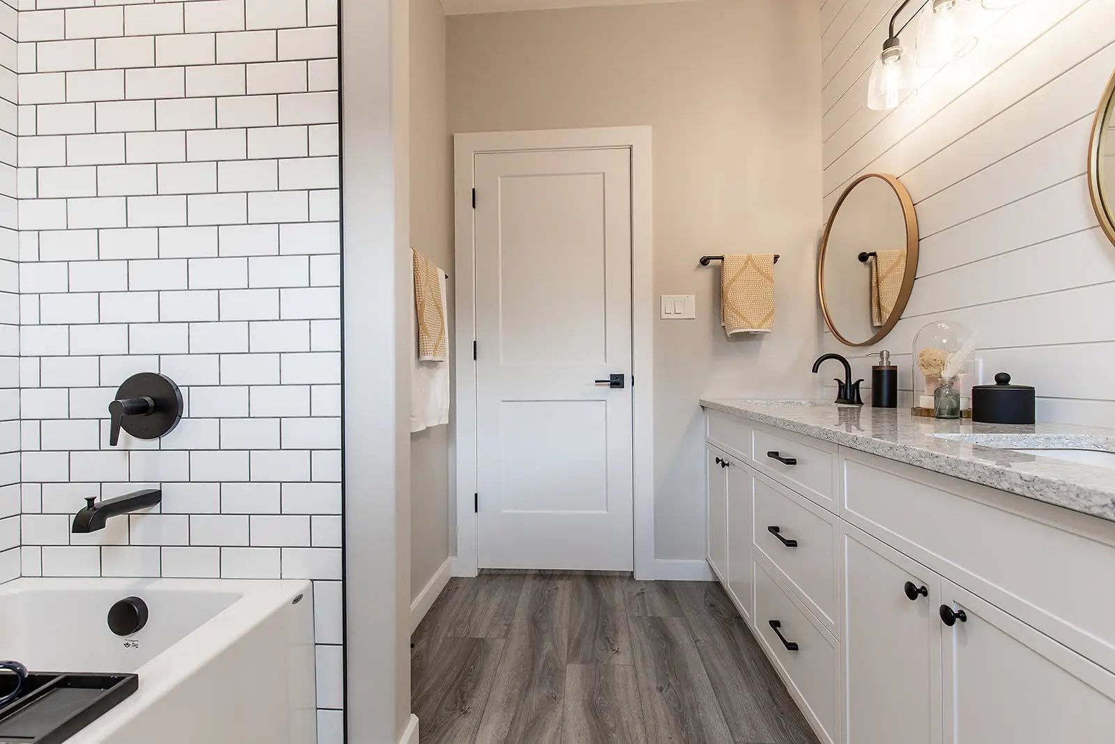 Modern bathroom with white subway tile, wood flooring, and black fixtures. Features a large mirror, double sinks, and bright, minimalist decor.