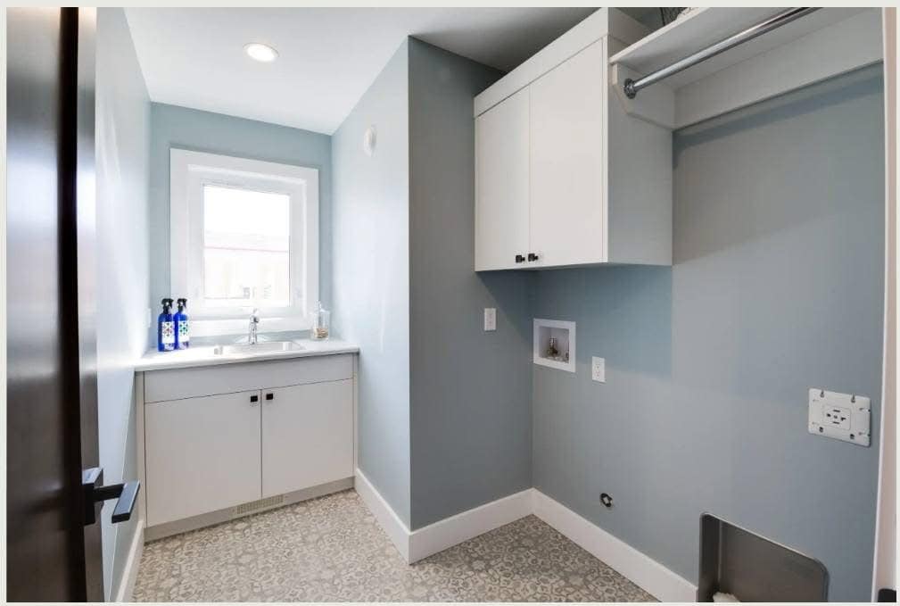 Modern laundry room with light blue walls and patterned gray floor. White cabinets are above a washing machine space. A sink by the window offers brightness.