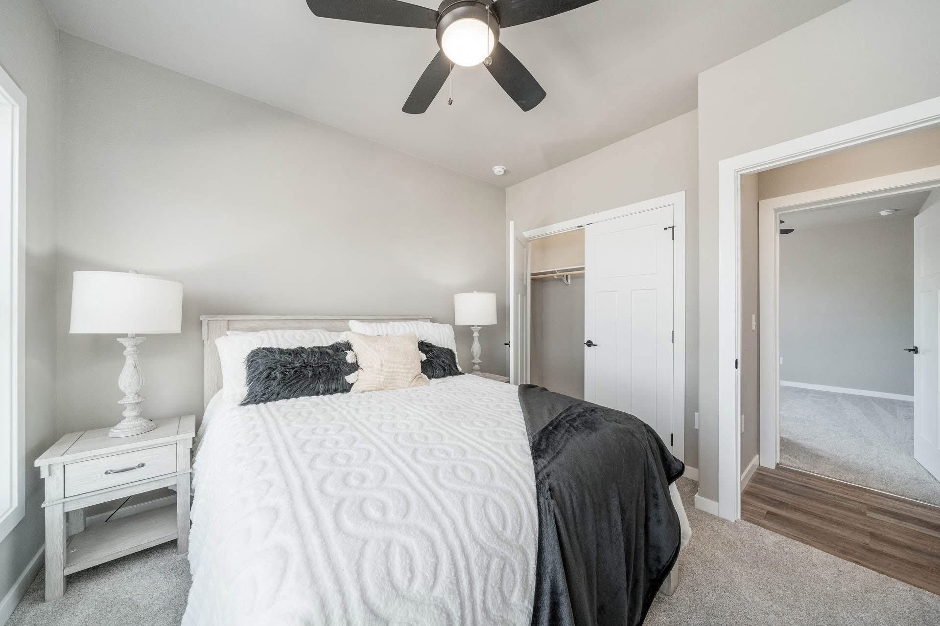 A bright bedroom features a large bed with white and gray bedding, flanked by two side tables with lamps. A ceiling fan is above, and an open closet door is on the right. The room feels serene and inviting.