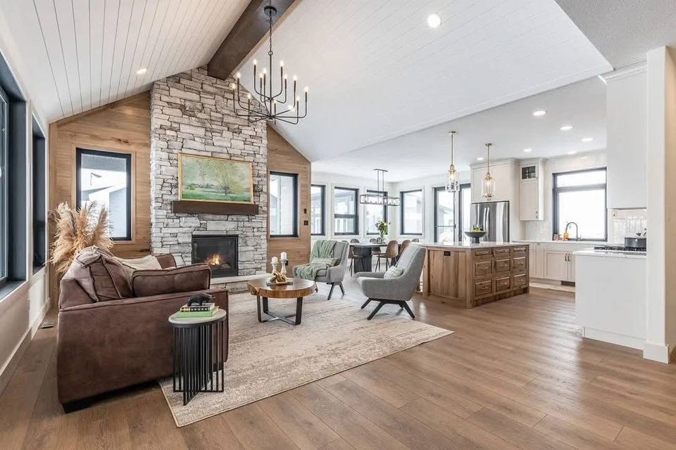 Spacious open-plan living area with a high ceiling and hardwood floors. Features a stone fireplace, cozy seating, a modern kitchen, and dining area.