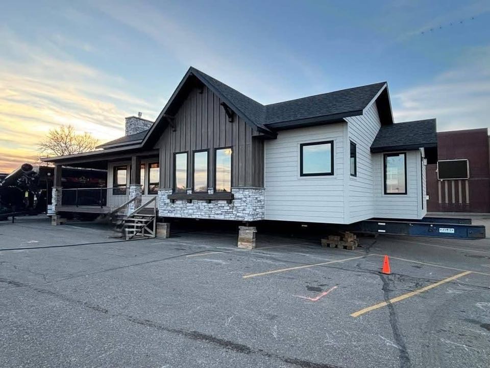 A prefabricated house on a platform with grey siding and a gabled roof sits in a parking lot. The sky is a blend of blue and orange at sunset.