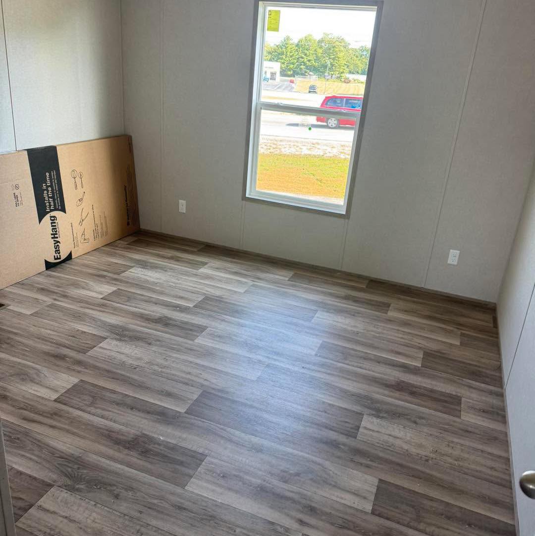 Empty room with gray wood vinyl flooring, white walls, and a single window letting in natural light. A large cardboard box leans against the wall.