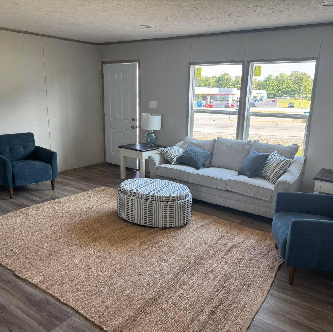 A cozy living room with a gray sofa, blue armchairs, a striped ottoman, and a large beige rug. Light streams through two large windows, creating a bright atmosphere.