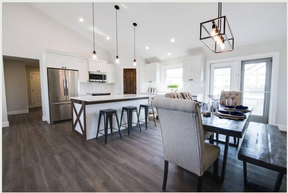 Spacious, modern kitchen and dining area with white cabinets, stainless steel appliances, an island with barstools, and a large wooden table under chic lighting.