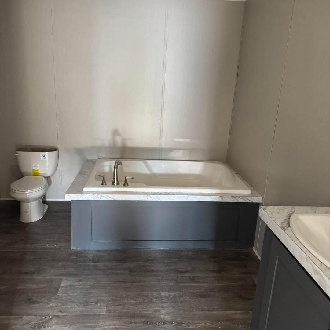 Modern bathroom featuring a white bathtub with silver faucets, adjacent to a toilet. Dark wood flooring adds warmth, and gray paneling creates a sleek look.