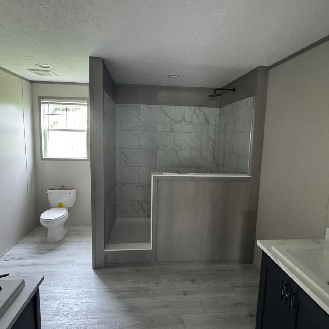 A modern bathroom with a marble-tiled open shower, white toilet under a window, and light wood flooring. The space appears clean and minimalist.