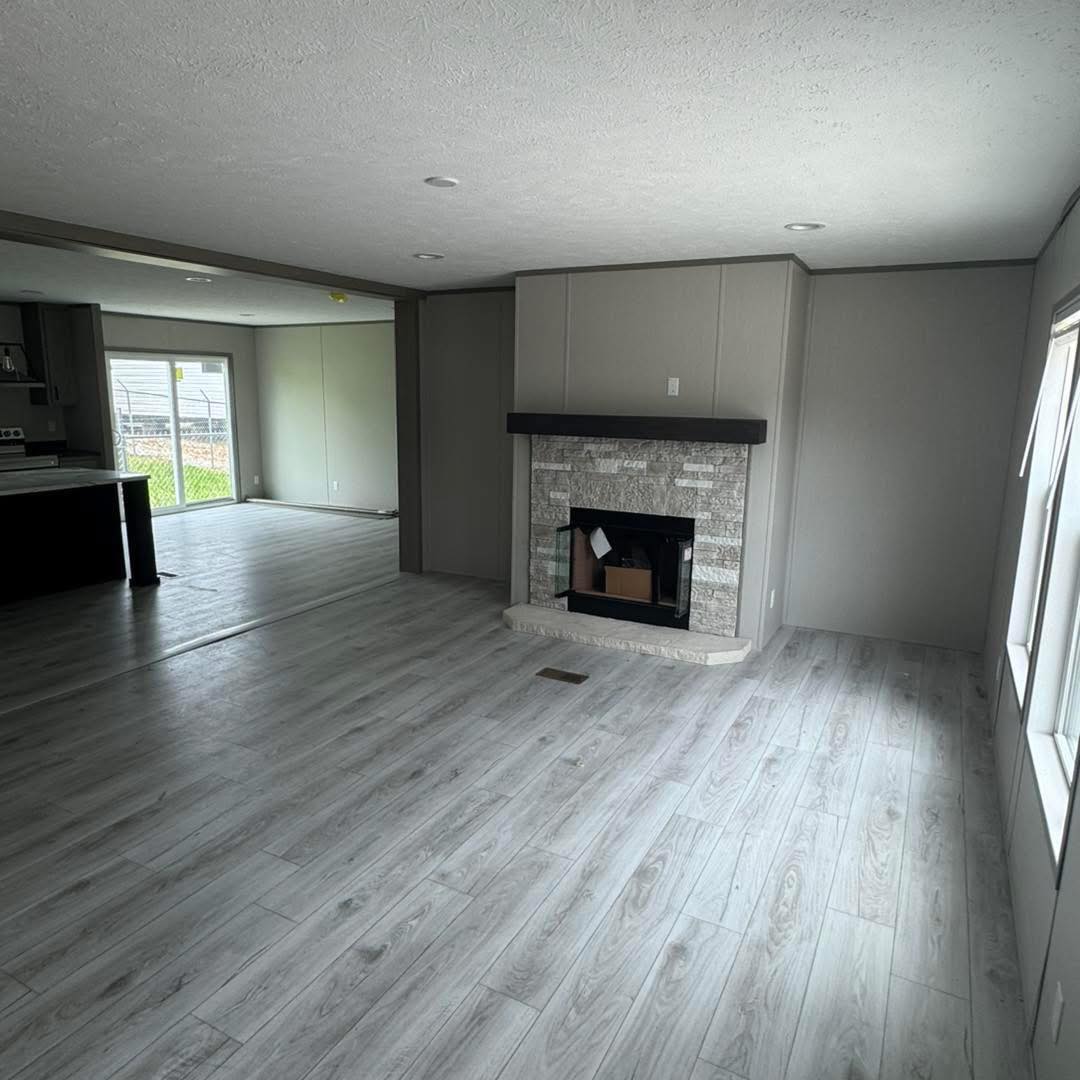 Spacious living room with light gray wood flooring, featuring a modern stone fireplace. Open layout leads to a kitchen with large windows and natural light.