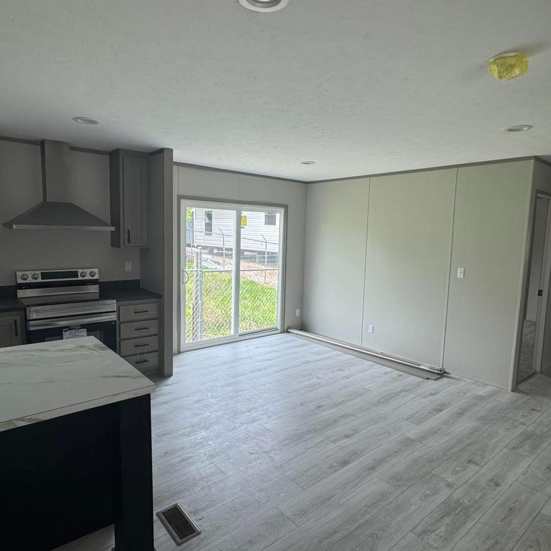 A bright, empty room with light wooden flooring. A modern kitchen, gray cabinets, and an oven are to the left. Sliding glass doors lead outside.