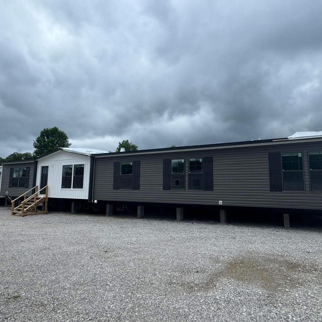 A long, dark gray mobile home with a white add-on section is parked on gravel, beneath an overcast sky, conveying a calm and somber mood.