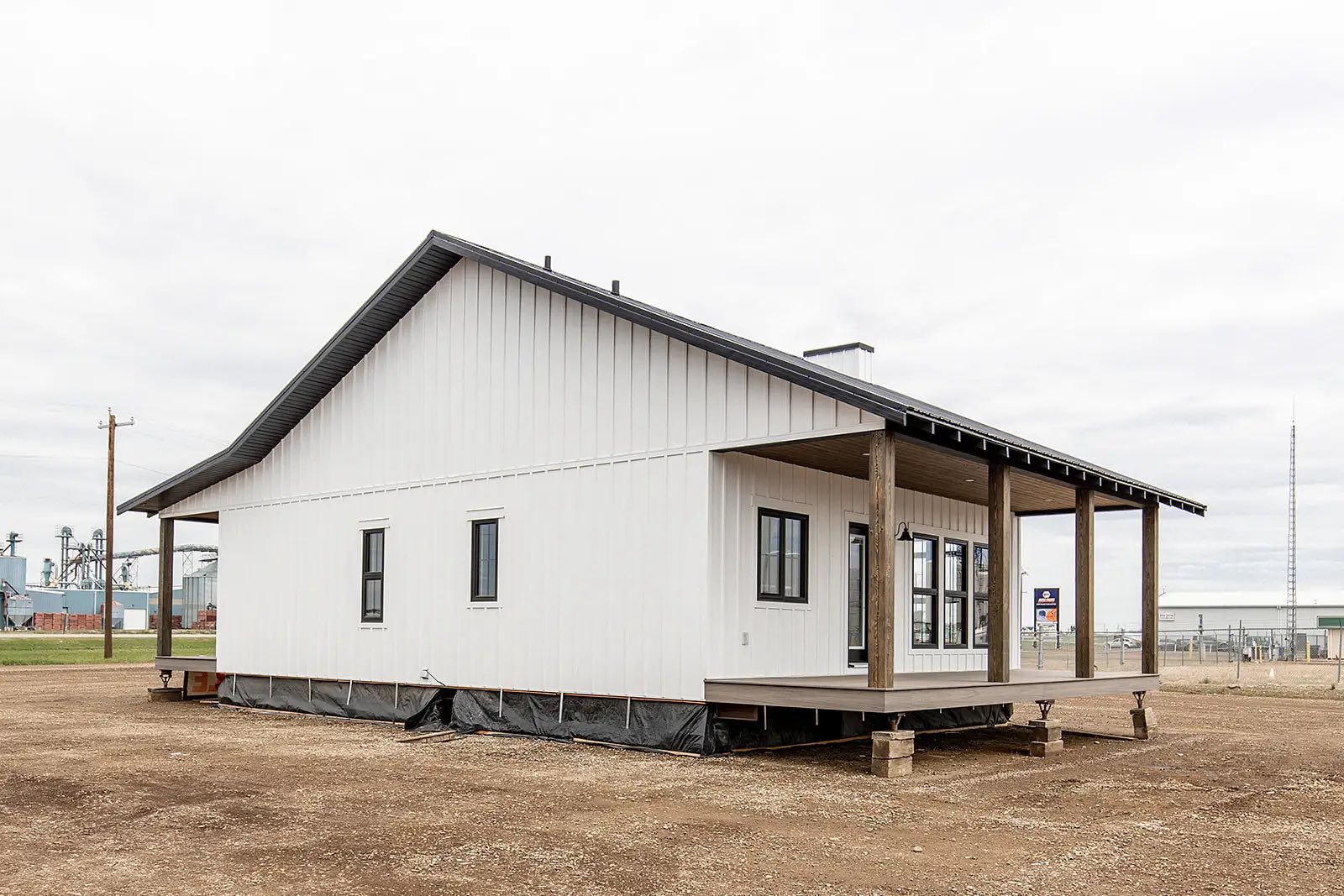 Single-story, modern white house with black trim and sloped roof. It features a wraparound porch with wooden pillars, set on a dirt plot under a cloudy sky.