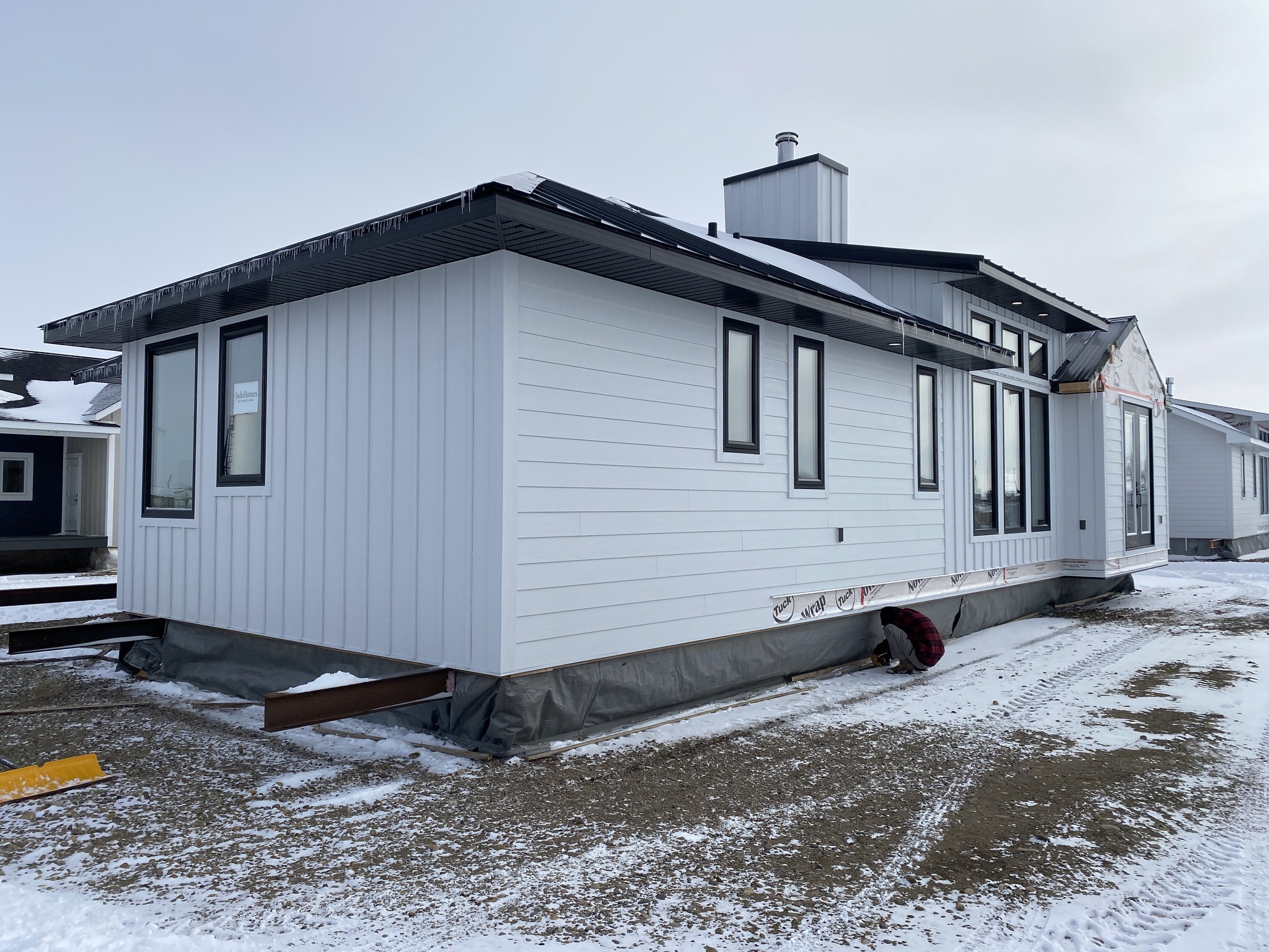 A partially constructed modular home sits on a snowy, dirt-covered lot. The exterior is light gray with black-framed windows, conveying a cold, barren atmosphere.