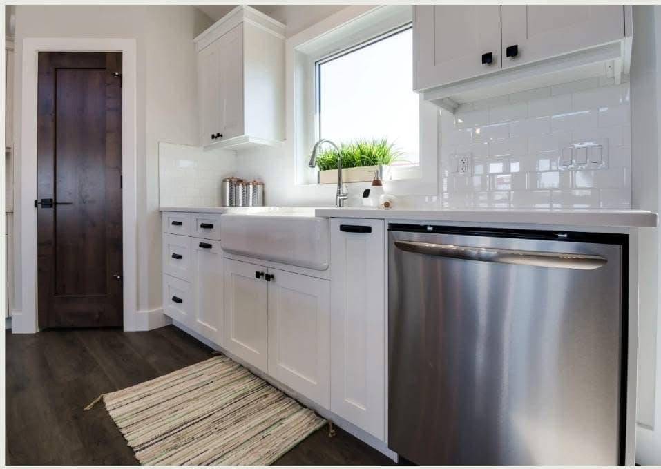 Modern kitchen with white cabinets, farmhouse sink, and stainless steel dishwasher. Sunlit window with potted plant; dark wood floor and door.