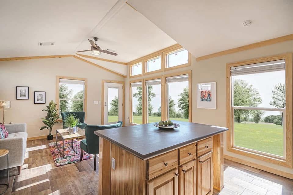Bright, modern living area with large windows overlooking a green landscape. Features a kitchen island, cozy seating, plants, and framed art.