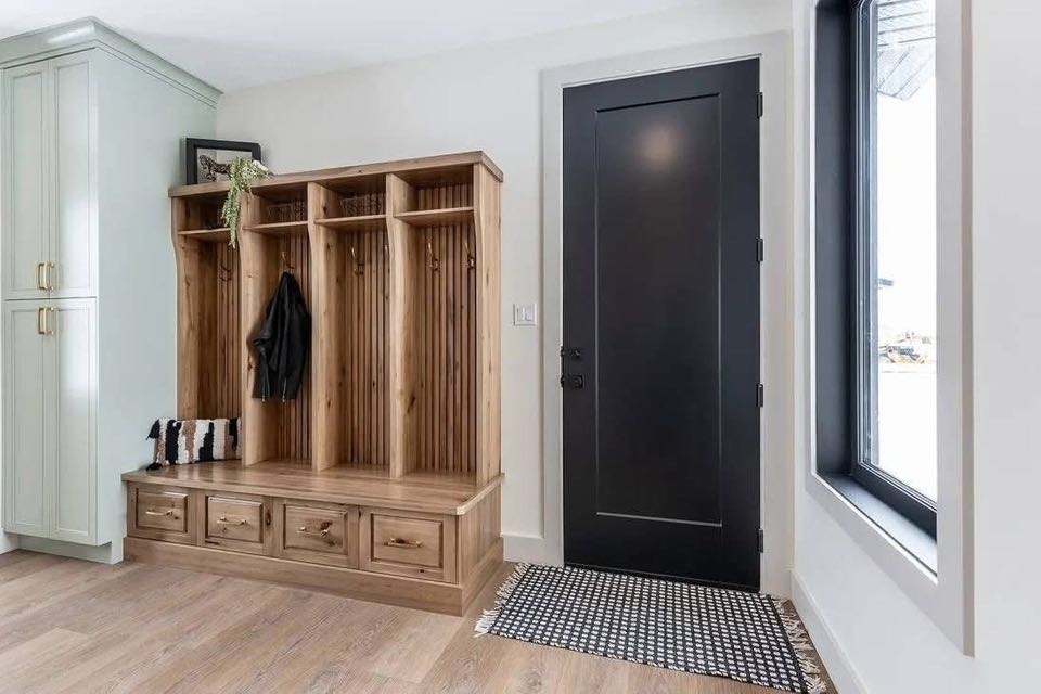 Modern mudroom with a light wooden organizer featuring hooks, shelves, and drawers. A black door and a window provide contrast. Neatly arranged, cozy vibe.