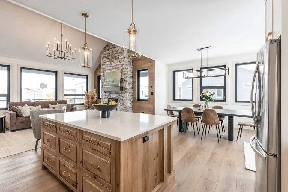 Modern open-concept kitchen and dining area with wooden floors. Features a large island, pendant lights, stone accent wall, and cozy seating, evoking warmth and style.