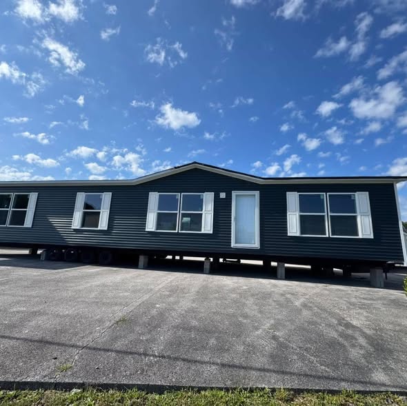 A blue mobile home with white shutters sits elevated on a concrete surface under a bright, partly cloudy sky, conveying a sense of openness and potential.