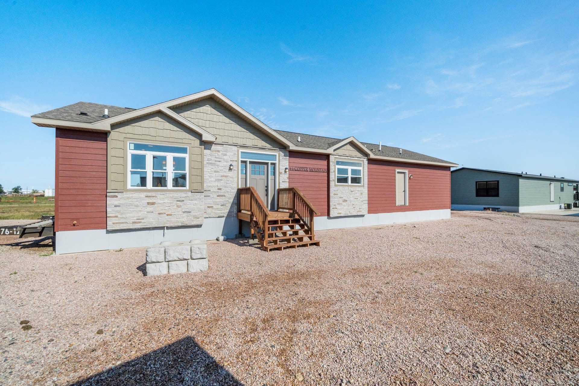 Single-story, beige and red manufactured home with stone accents and a small wooden porch. It's set on a gravel lot under a clear blue sky.