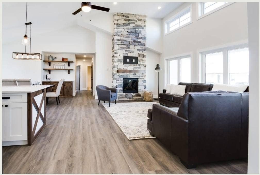 Modern open-concept living room with high ceilings, featuring a stone fireplace, large windows, wooden floors, dark leather sofas, and a kitchen with pendant lighting.