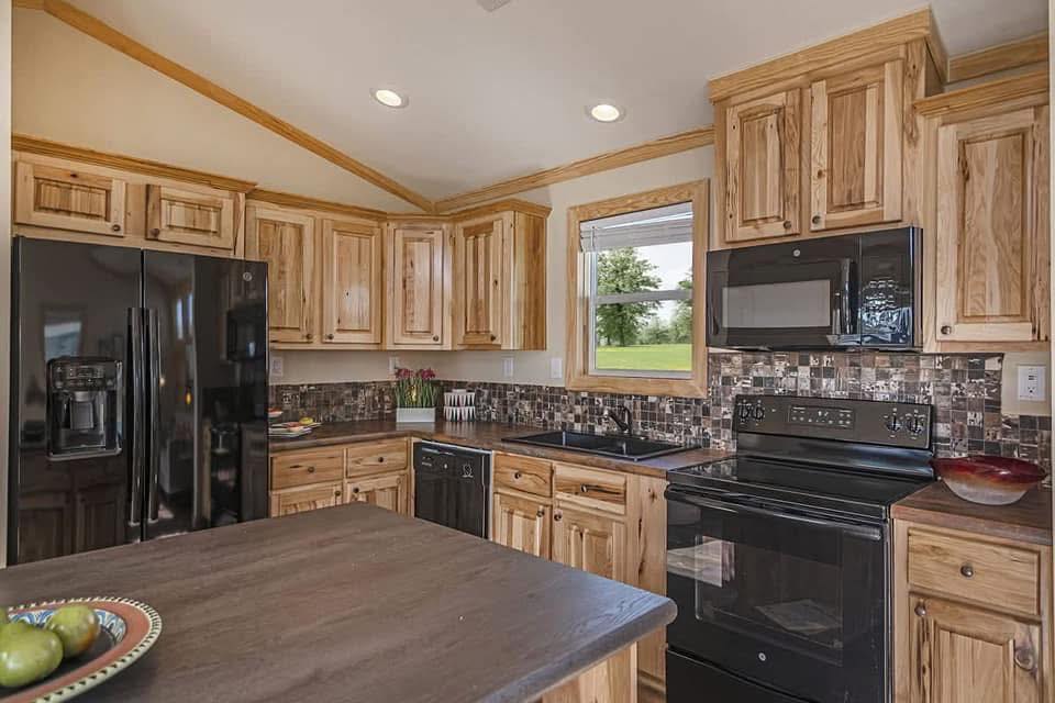Rustic kitchen with wood cabinets, black appliances, and a dark countertop. A window offers a view of greenery, adding warmth and charm to the space.