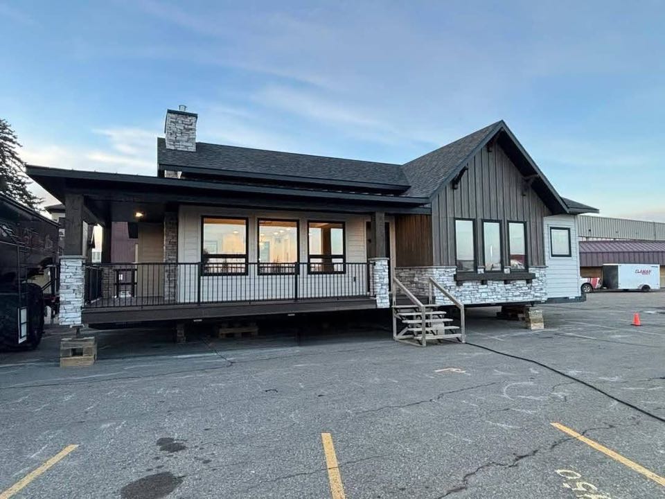 A modern, single-story modular home with dark wood paneling and stone accents, elevated on blocks in a parking lot, under a clear evening sky.