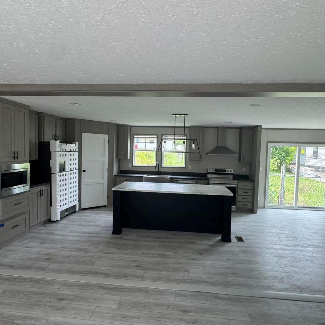 Modern kitchen with gray cabinets, a large island with a white countertop, stainless steel appliances, and light wood flooring. Bright natural light from large windows.
