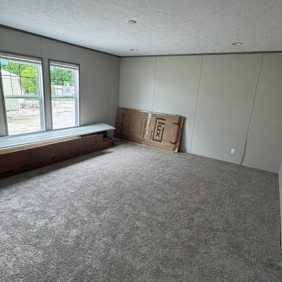 Empty room with gray carpet, light gray walls, and two large windows showing a view of trees. Cardboard boxes are stacked against one wall. Calm atmosphere.