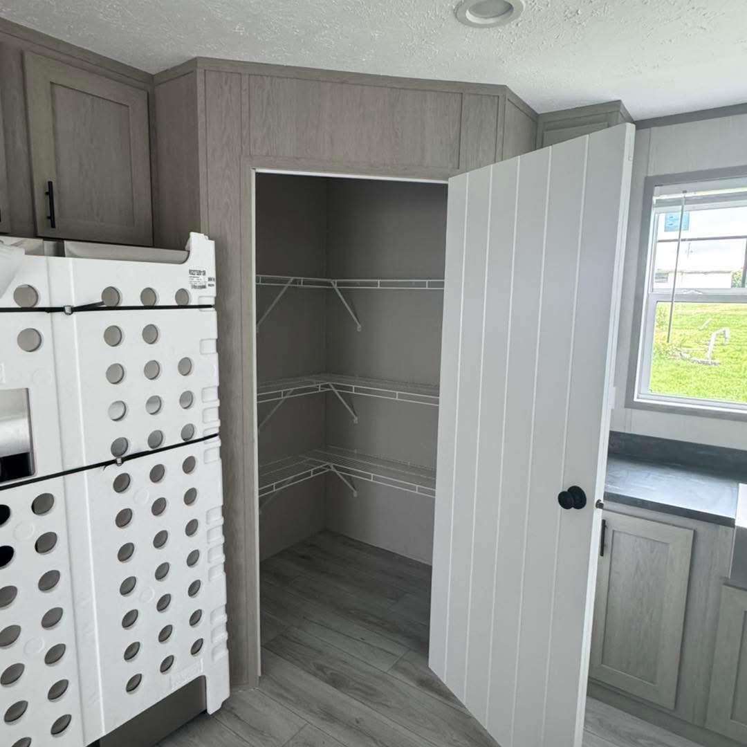 A small pantry with open white door and empty white metal shelves. Nearby is a stack of large, wrapped boxes. A window provides natural light, showing a grassy lawn outside.