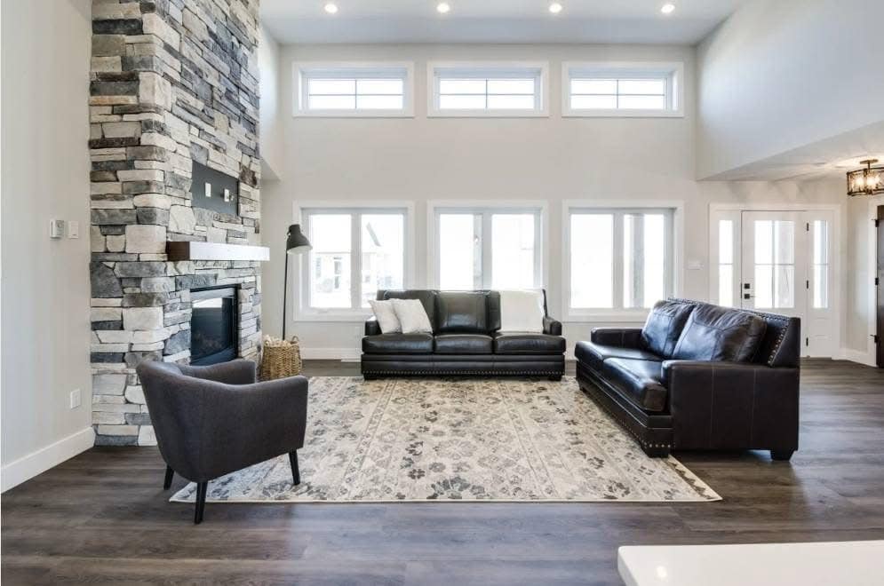 Modern living room with high ceilings and large windows. Features a stone fireplace, dark leather sofas, a gray armchair, and a patterned rug. Bright and inviting.