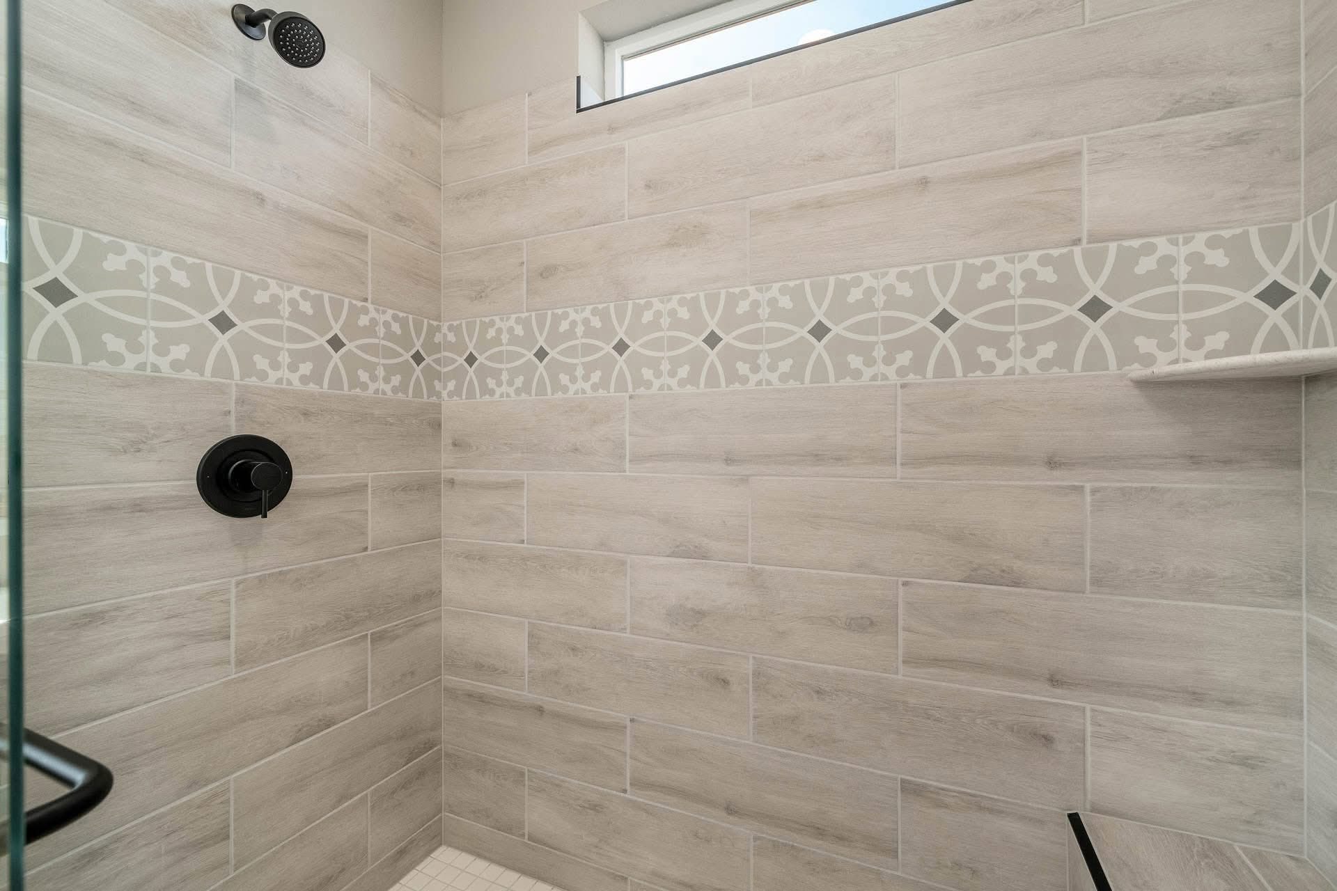 Shower with beige tiles, decorative border featuring geometric patterns. Black fixtures, small window above for natural light, creating a modern look.