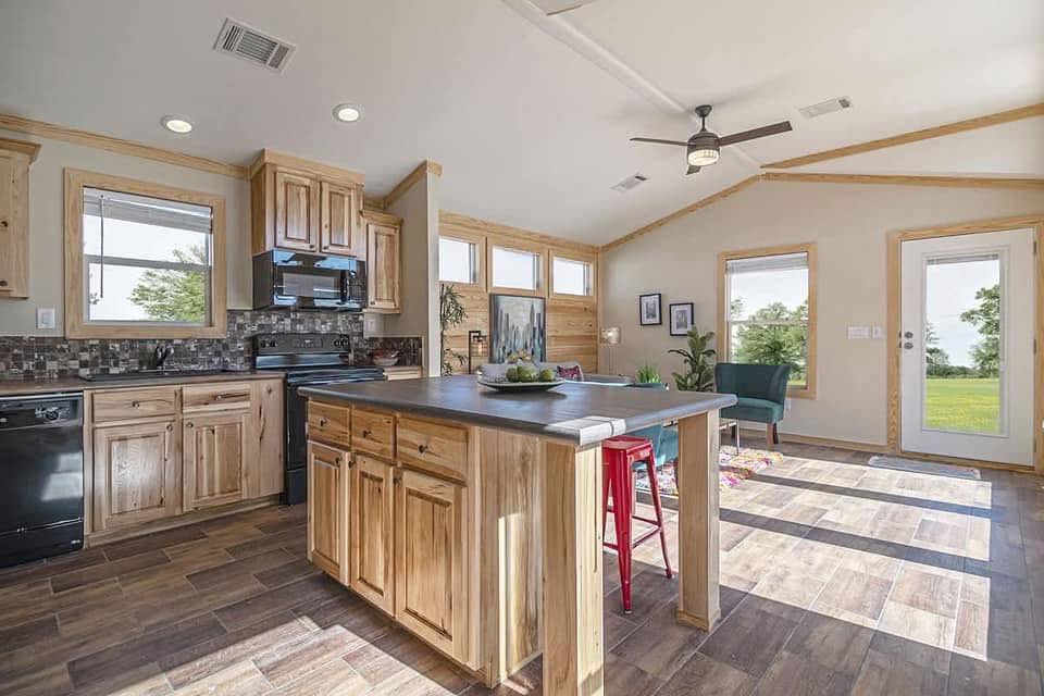 Bright, cozy kitchen-living space with light wood cabinets, central island, red stools, and large windows. Room is sunny, modern, and inviting.