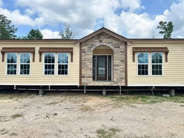 A beige modular home with a stone entrance, six large windows, and a dark brown door. It sits on a gravel lot under a partly cloudy sky.