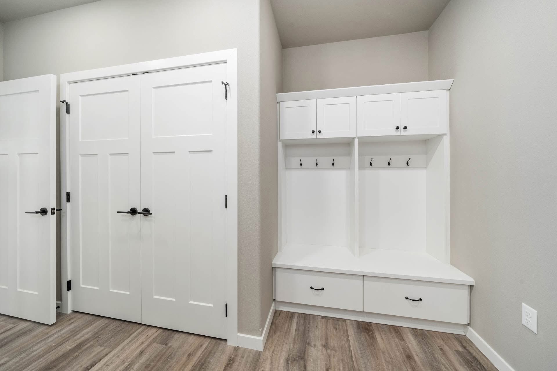 Modern mudroom with white cabinets and wood flooring. Includes a storage bench with drawers, hooks above for hanging, and closed closet doors.