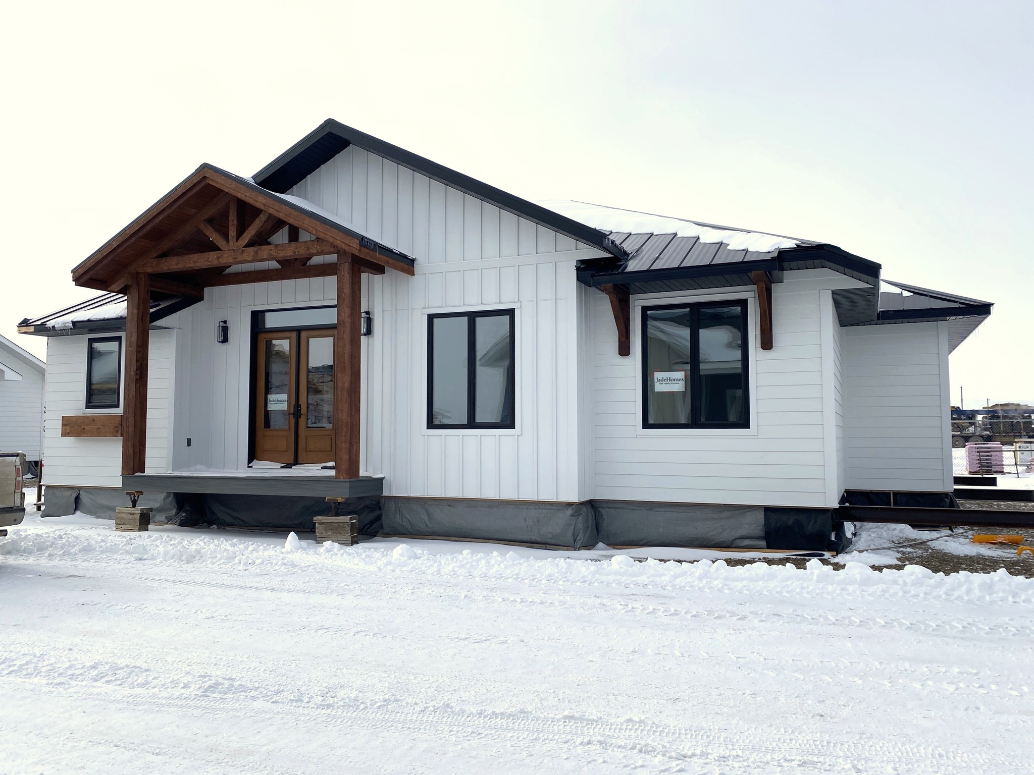 A small, modern white house with dark windows and a wooden porch stands against a snowy landscape under a bright sky, exuding a cozy and inviting feel.