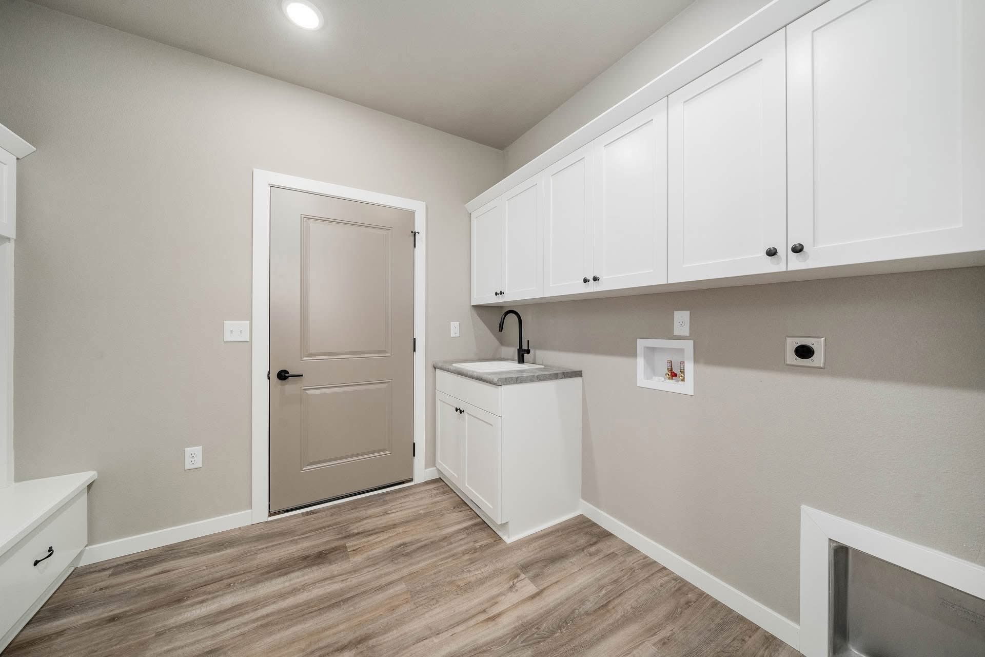 A modern laundry room with light wood flooring. White cabinets and a gray countertop with a black faucet are beside a utility sink and beige door.