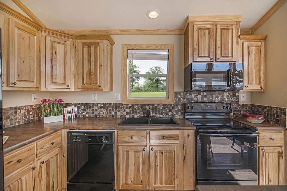 A cozy kitchen with natural wood cabinets, a black stove and microwave, a patterned backsplash, and a window showing a green landscape outside.