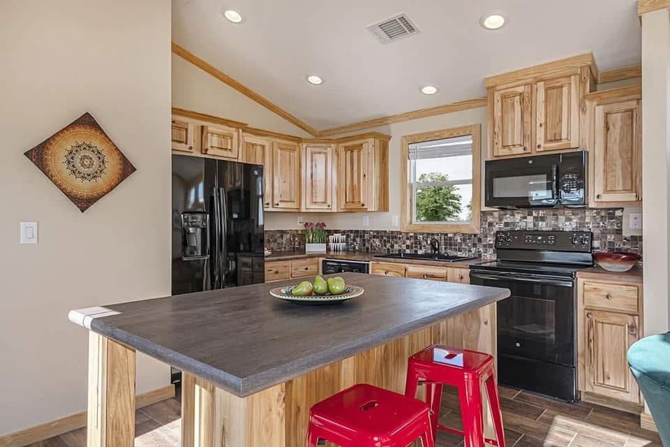 A cozy kitchen with natural wood cabinets, black appliances, and mosaic tile backsplash. A dark island with green apples and red stools adds contrast.